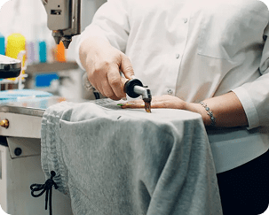 Person using a handheld tool on a piece of gray fabric, possibly for sewing or tailoring, in a workspace with sewing equipment.