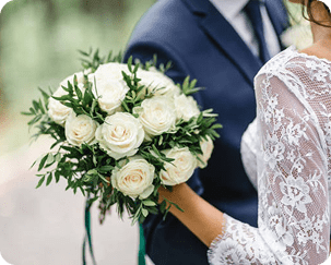 Bride holding a bouquet of white roses, standing next to a groom in a navy suit. Both are partially visible, with focus on the flowers and lace detail of the bride's sleeve.