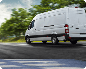 A white delivery van drives on a road with blurred green trees in the background, indicating motion.