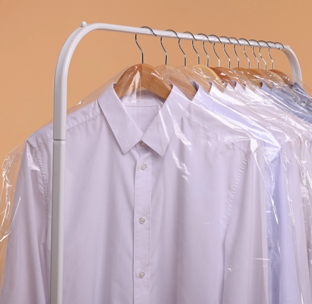 Several white dress shirts on wooden hangers are covered in plastic garment bags and hanging on a white clothing rack against a beige background.