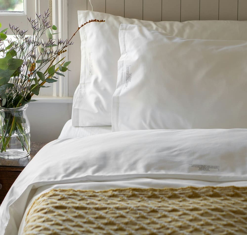 A neatly made bed with white pillows and sheets, a textured beige blanket, and a glass vase of flowers on a wooden bedside table by a window.