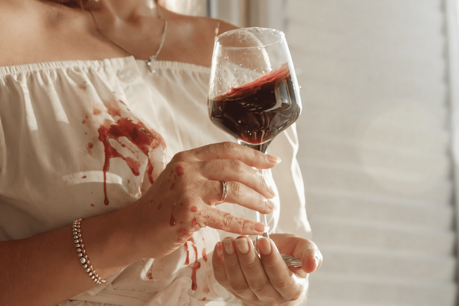 A person holding a glass of red wine with wine spilled on their white dress and hand.